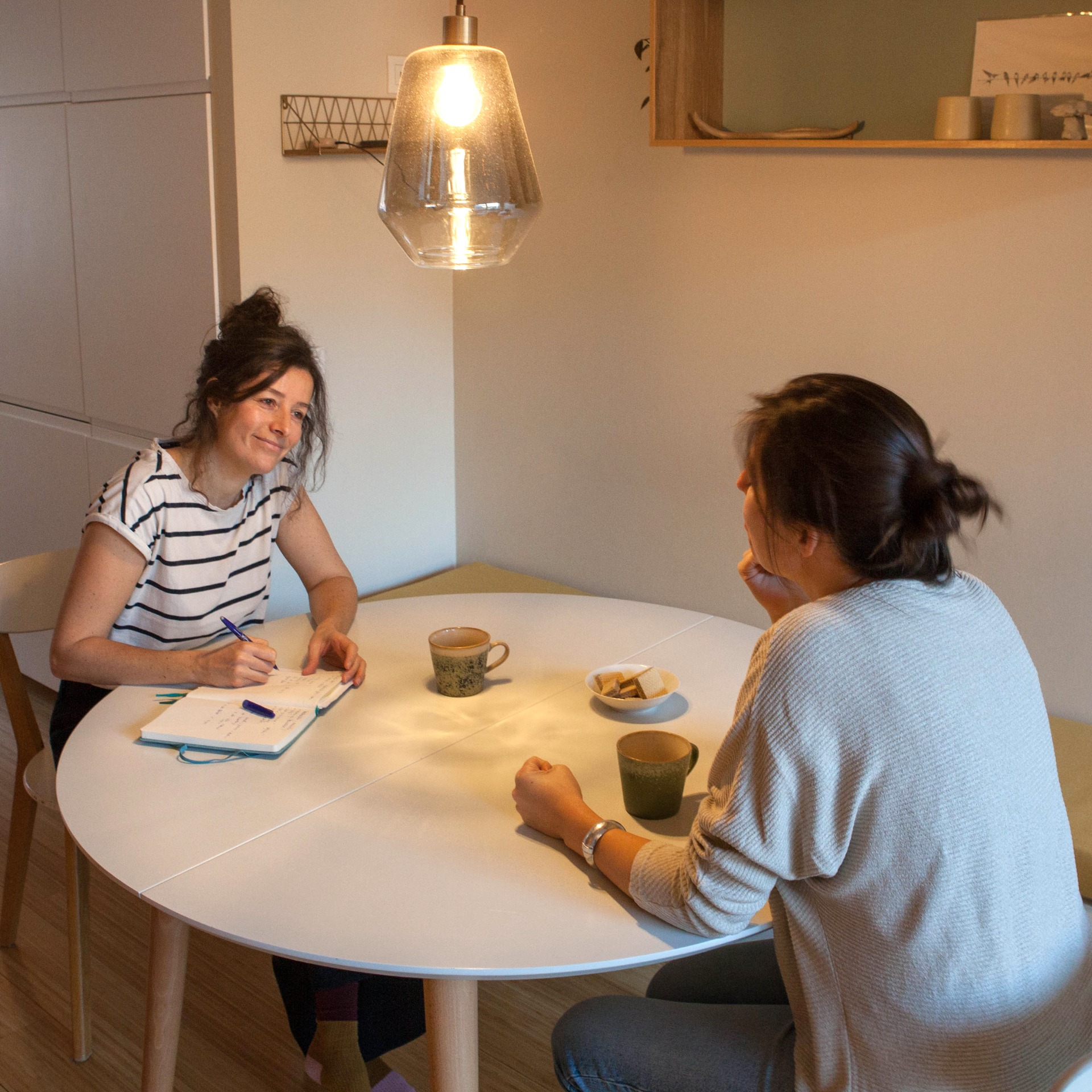 Twee vrouwen zitten aan een ronde tafel, schrijven in een notitieboek onder warm hanglamplicht.